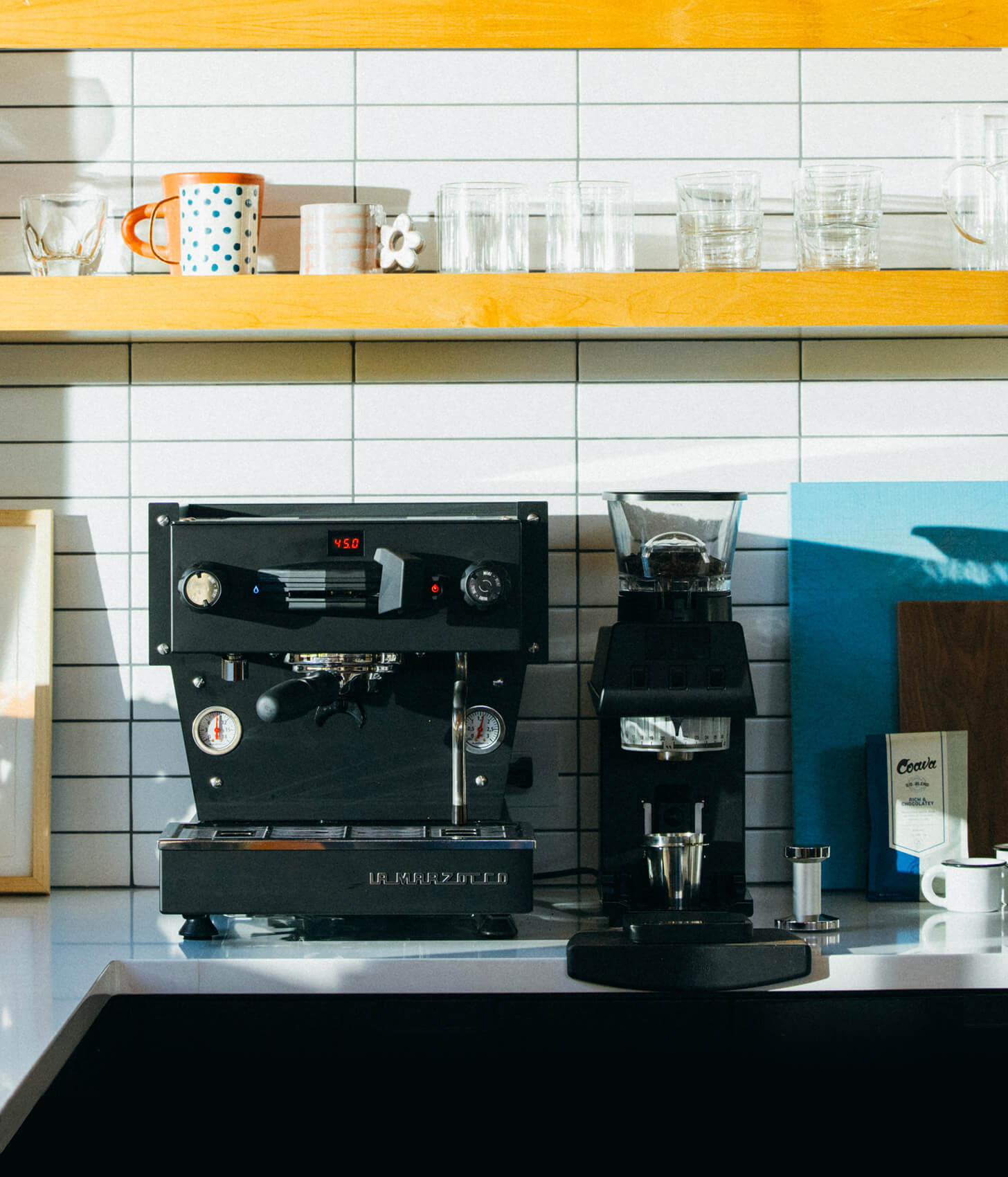 Sort La Marzocco Linea Mini R espressomaskine og sort Pico kaffekværn på hvid køkkenbænk med gule hylder.